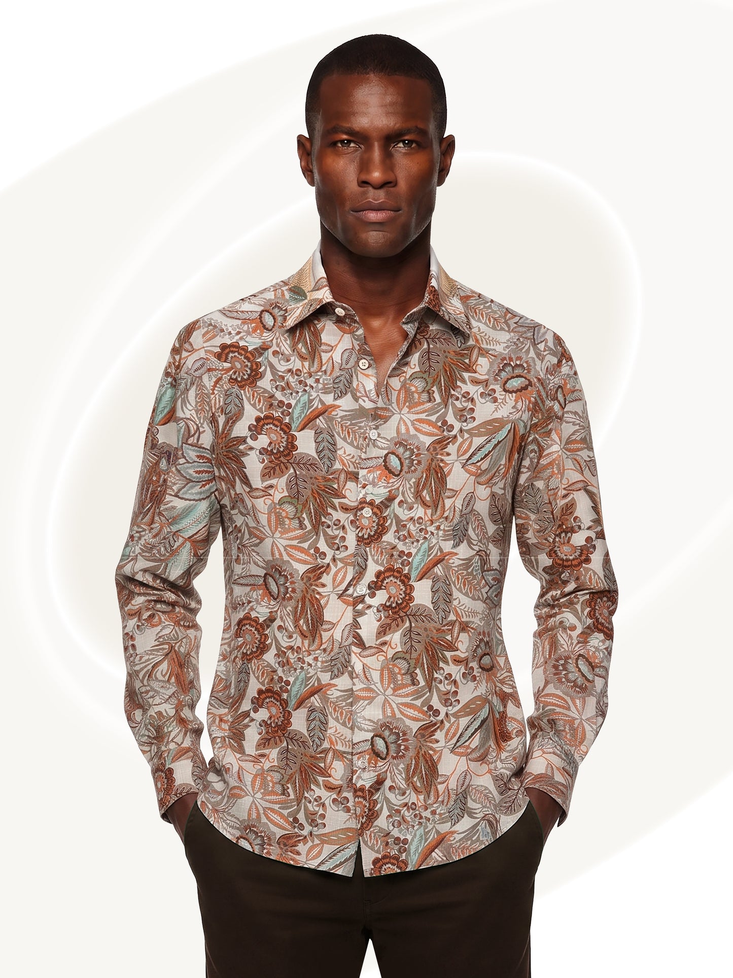 A man wearing a long-sleeve button-up shirt with an intricate botanical and paisley pattern in brown, beige, and muted blue, paired with dark brown trousers, set against a light off-white background with subtle circular graphic elements.