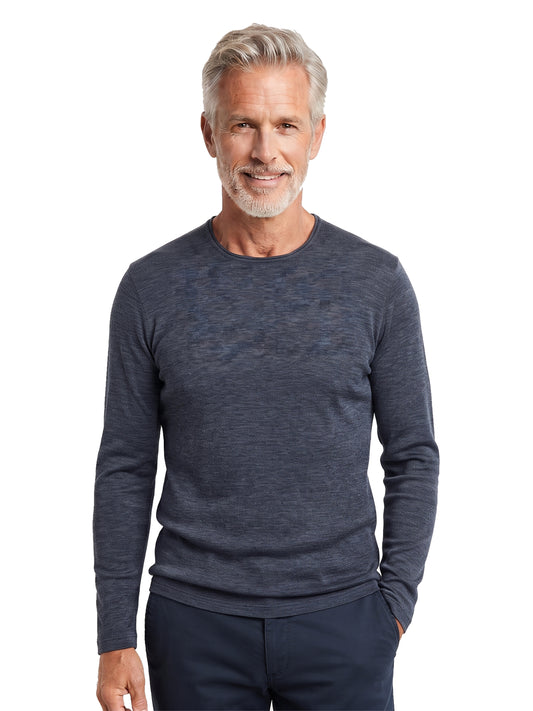 A smiling older man with gray hair and a beard wearing a dark heathered blue long-sleeve crewneck sweater and dark pants, standing against a plain white background.