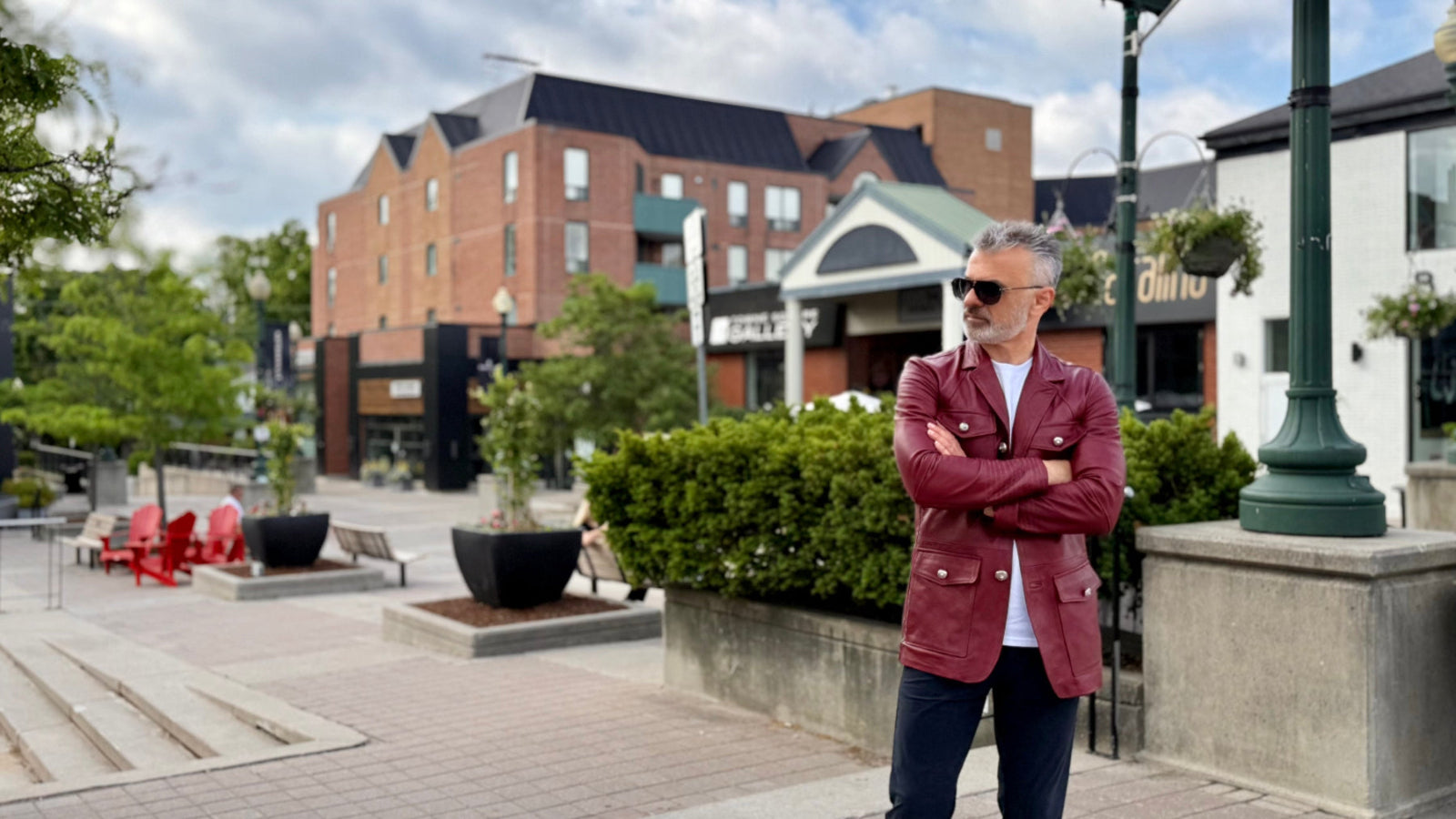 Man in a red coat standing in an outdoor urban setting with buildings and greenery.