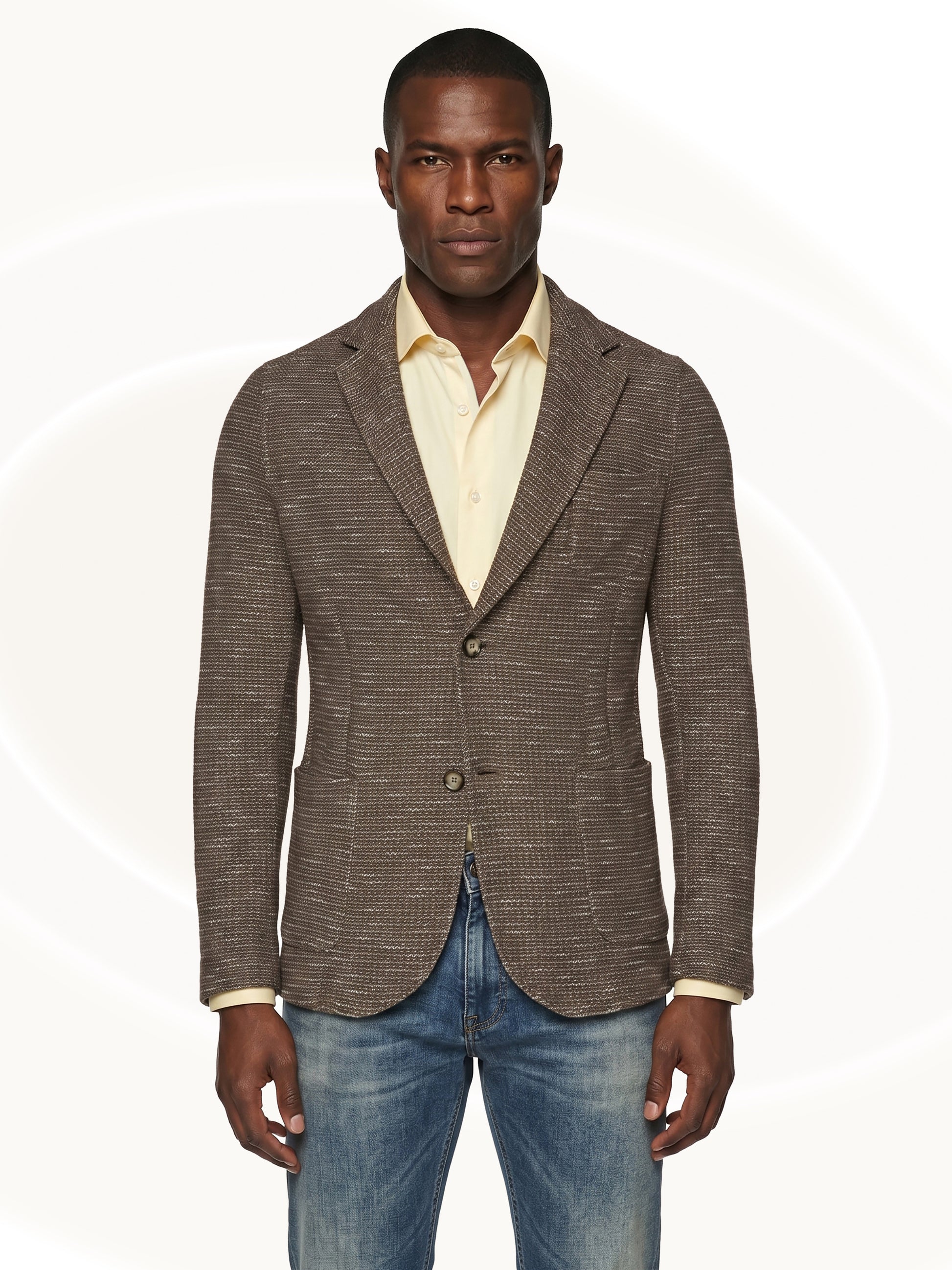 Man wearing a brown fitted blazer over a yellow shirt and blue jeans on a white background
