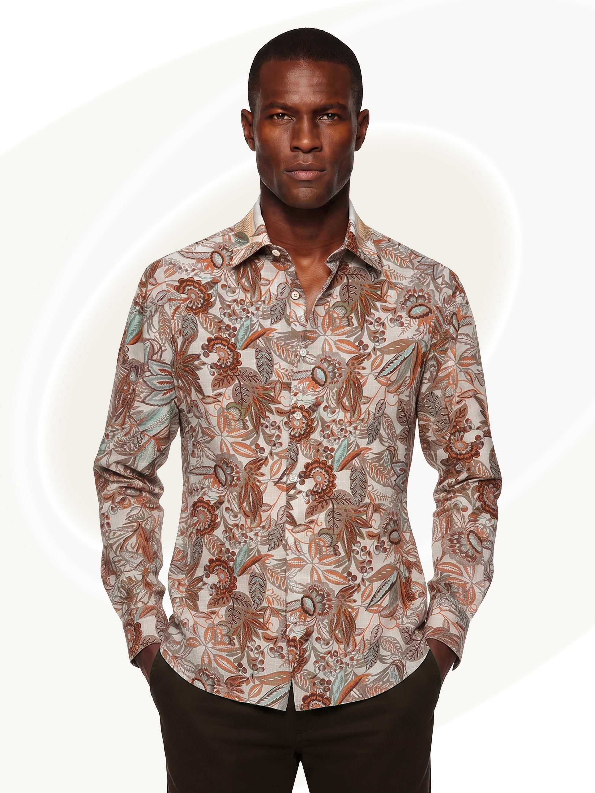 A man wearing a long-sleeve button-up shirt with an intricate botanical and paisley pattern in brown, beige, and muted blue, paired with dark brown trousers, set against a light off-white background with subtle circular graphic elements.